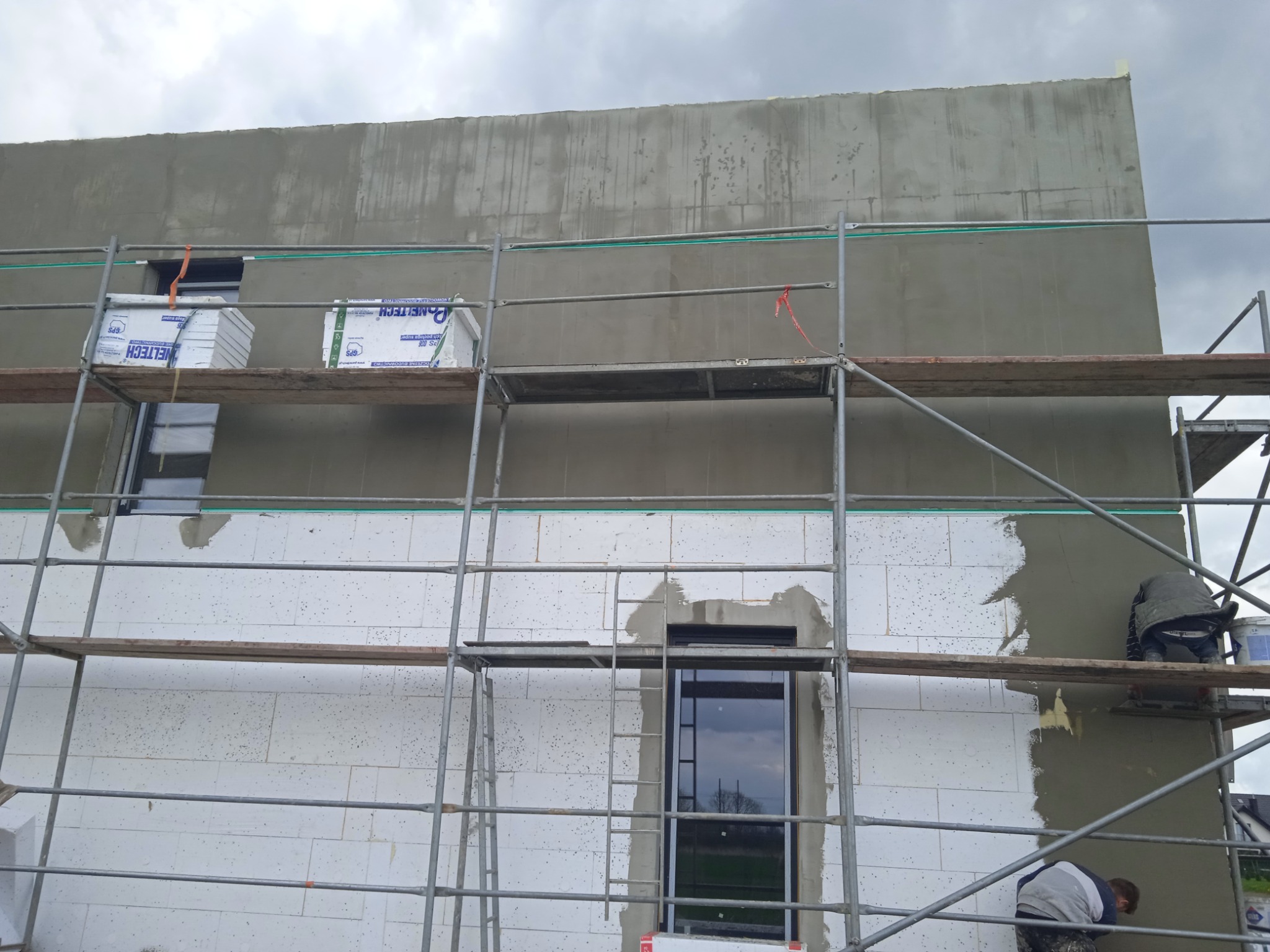 Ocieplanie budynku z białych bloczków, fragment ściany pokryty szarym tynkiem, widoczne rusztowanie i pracownicy podczas nakładania tynku.
