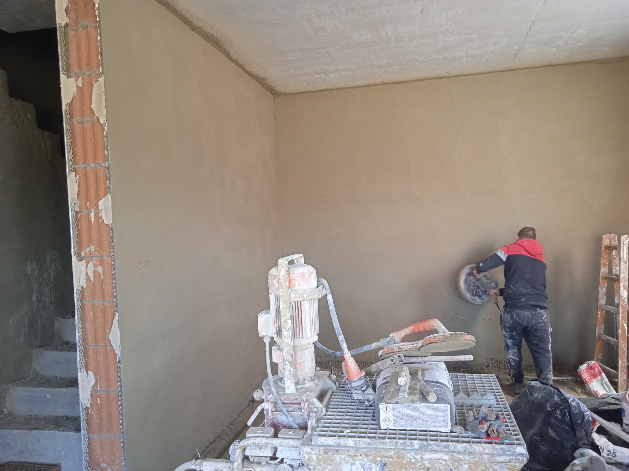 Pracownik wyrównuje świeżo nałożony tynk na ścianie za pomocą szlifierki, widoczna maszyna do tynkowania, schody w tle.