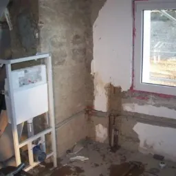 Surowy stan łazienki w trakcie remontu: stelaż podtynkowy WC, odsłonięte rury, ściany z resztkami tynku i nowym oknem.