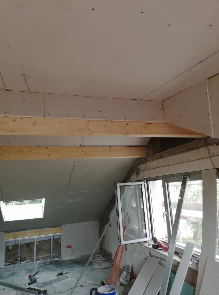 Montaż płyt gipsowo-kartonowych na poddaszu z drewnianą konstrukcją nośną. Widoczne okno dachowe i narzędzia pracy.