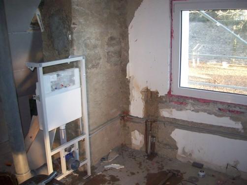 Surowy stan łazienki w trakcie remontu: stelaż podtynkowy WC, odsłonięte rury, ściany z resztkami tynku i nowym oknem.