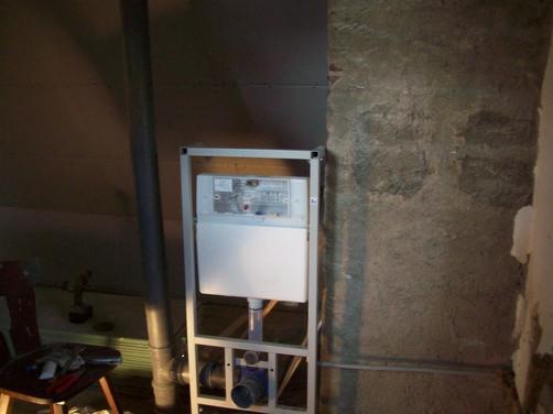 Stelaż podtynkowy WC Geberit Duofix z widocznymi elementami instalacji wodno-kanalizacyjnej, w trakcie montażu przy surowej, betonowej ścianie. Obok, na krześle leżą narzędzia.