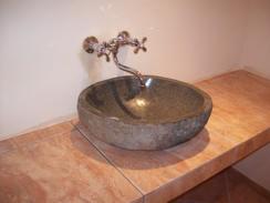 Umywalka z szarego kamienia o nieregularnym kształcie, osadzona na blacie z płytek imitujących marmur. Bateria chromowana, dwu-uchwytowa w stylu retro. Ściana w tle w odcieniu kremowym.