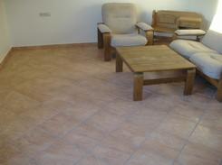 Jasne wnętrze pokoju z brązową, wzorzystą podłogą z płytek, drewnianym stolikiem kawowym i częściowo widocznym jasnym narożnikiem.