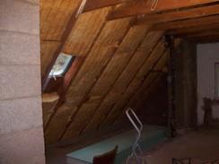 Poddasze w trakcie docieplania wełną mineralną, widoczna skośna konstrukcja dachu z zamontowanymi panelami izolacyjnymi, małe okno dachowe, drabina oparta o ścianę, surowy stan wykończenia wnętrza.