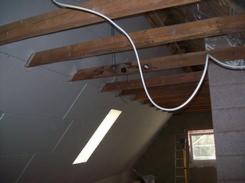 Skośny strych w trakcie docieplania, widoczne drewniane belki stropowe, biała instalacja elektryczna oraz okno dachowe z silnym refleksem słonecznym. Część ściany pokryta szarym materiałem...