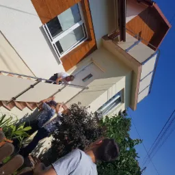 Montaż balustrady balkonowej z wypełnieniem z paneli na tle elewacji domu, widoczne fragmenty drewnianej podbitki dachu, okna i roślinność.