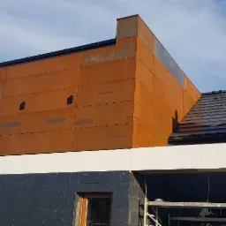 Nowoczesny dom z elewacją z paneli imitujących rdzę, widok z dołu na fragment budynku z oknem i fragmentem dachu z czarnej dachówki.