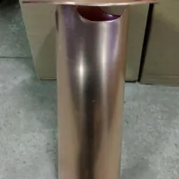 Miedziany wywietrznik dachowy z daszkiem w kształcie stożka, na szarym podłożu, w tle kartonowe pudła. Metaliczny połysk.