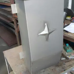 Szara, metalowa skrzynka z ozdobną, spawaną gwiazdą na bocznej ściance, stojąca na stole warsztatowym w zakładzie produkcyjnym. Widoczne ślady obróbki.