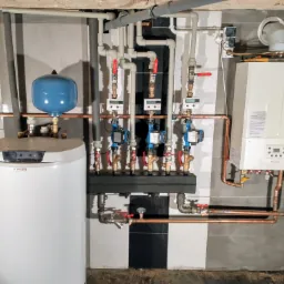 Nowoczesna kotłownia z białym zasobnikiem ciepłej wody Drazice, piecem gazowym Immergas, rozdzielaczem i pompami obiegowymi na tle biało-szarych płytek.