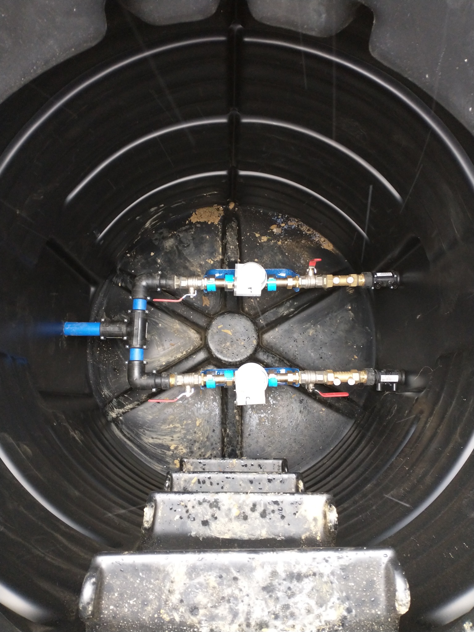 Wnętrze czarnego zbiornika retencyjnego z zainstalowanymi dwoma rzędami rur i zaworów, widok z góry, lekko zabrudzone.