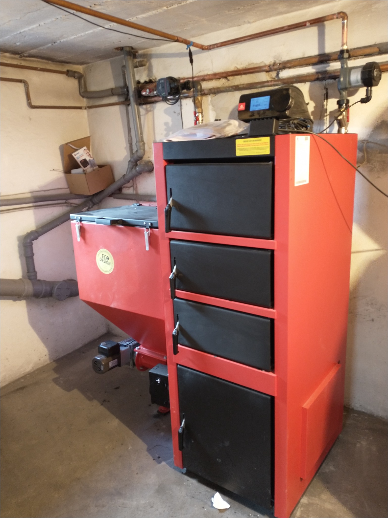 Czerwony piec na paliwo stałe z czarnymi drzwiczkami w piwnicy, widoczne rury instalacji grzewczej pod sufitem i zasobnik na paliwo z naklejką Eco Design