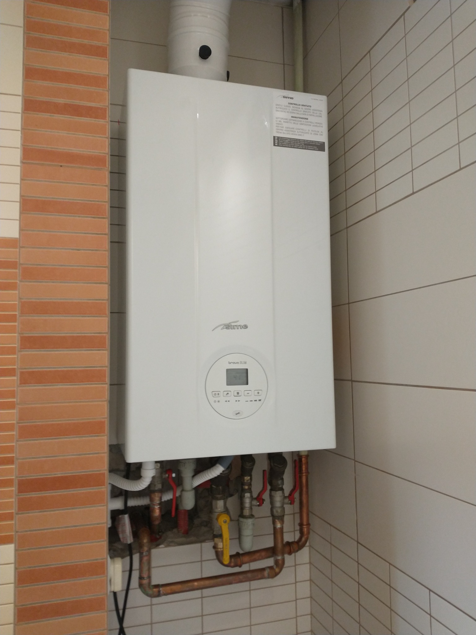 Nowy, biały piec gazowy marki Sime, zamontowany na ścianie z białych płytek, z widocznymi miedzianymi rurami i zaworami, zainstalowany w rogu pomieszczenia z kolumną z cegieł.