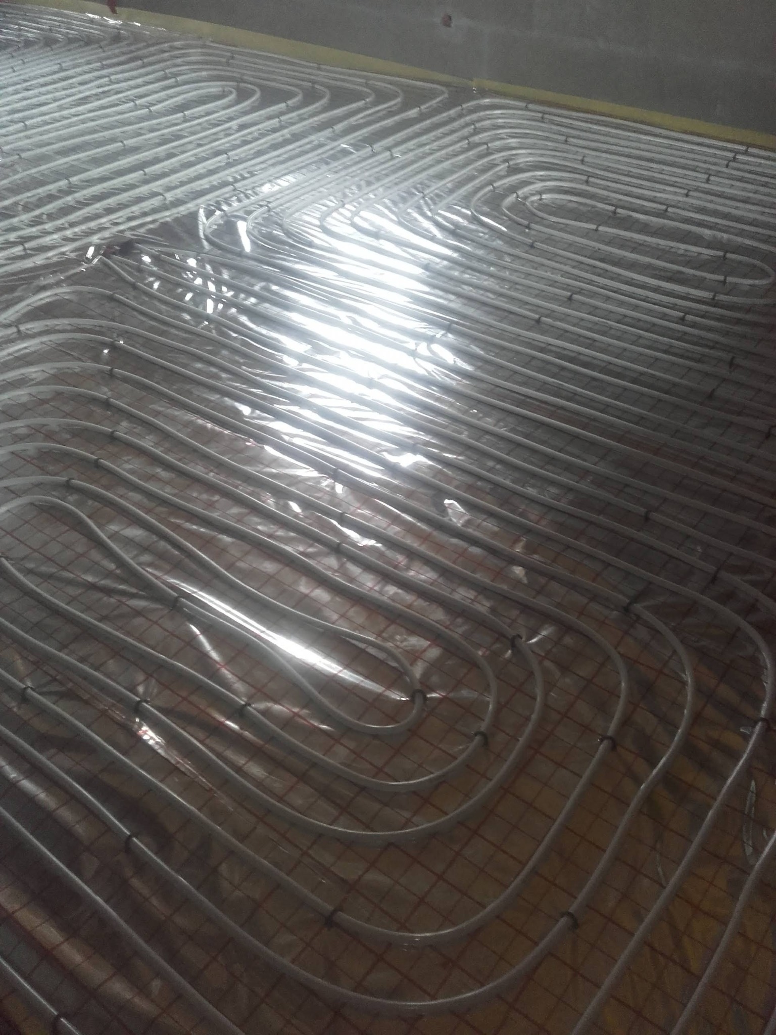 Białe rury ogrzewania podłogowego ułożone w pętle na folii odblaskowej z siatką, widoczne czarne zaciski mocujące.