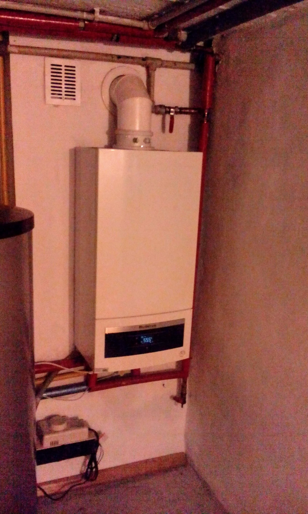 Biały piec gazowy marki Buderus zamontowany na czerwonym stelażu, z widocznymi rurami i przewodami, w pomieszczeniu o surowym wykończeniu.