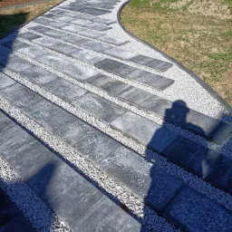 Ścieżka z prostokątnych płyt betonowych, wysypana białym grysem, wijąca się przez trawnik w ogrodzie, z widocznym cieniem osoby fotografującej.