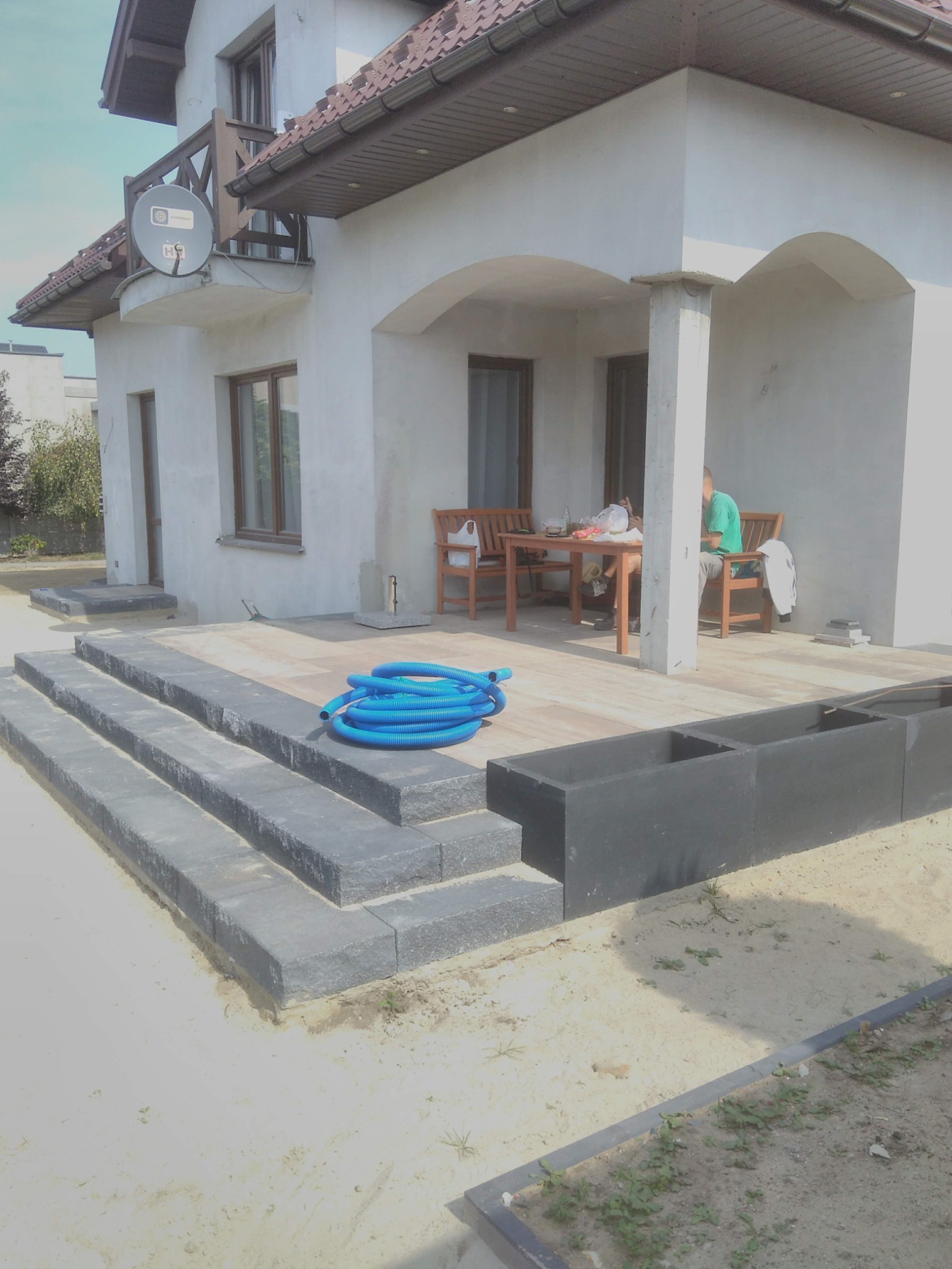 Nowe, grafitowe stopnie z kostki brukowej prowadzące na taras domu, z zwiniętym niebieskim wężem ogrodowym i donicami w tle.