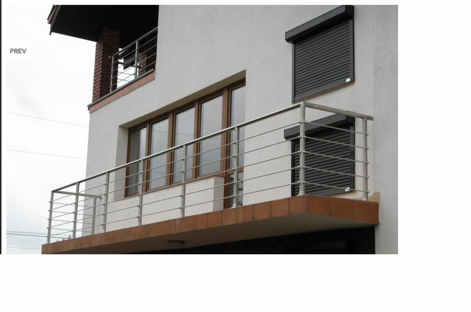 Balkon z nowoczesną balustradą ze stali nierdzewnej, z poziomymi prętami, na tle białej elewacji budynku z brązowymi oknami i roletami zewnętrznymi.