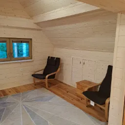 Jasny pokój na poddaszu wykończony drewnem, z dwoma fotelami z ciemną tapicerką, wzorzystym dywanem i oknem z widokiem na zieleń.