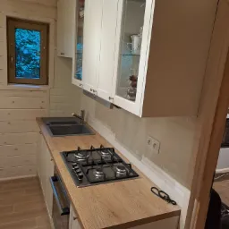 Drewniana kuchnia w stylu skandynawskim z białymi szafkami z przeszklonymi frontami, szarą płytą grzewczą i zlewozmywakiem, widok z boku na blat z jasnego drewna i okno z widokiem na las.