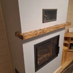 Szczegółowe ujęcie nowoczesnego kominka z czarną ramą i drewnianą półką o wyraźnym usłojeniu, zamontowanego w szarej ścianie z kratką wentylacyjną, w otoczeniu drewnianych ścian i mebli.