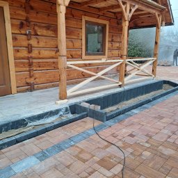 Suchodolski - Drewniany dom z bali w trakcie budowy tarasu z kostki brukowej. Widoczne elementy konstrukcyjne domu i częściowo ułożona kostka w odcieniach brązu i szarości.