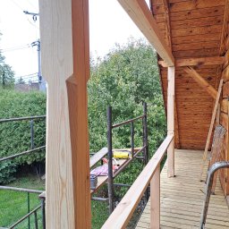 Suchodolski - Drewniany taras w trakcie budowy: widoczne deski podłogowe, balustrada, słupy nośne z charakterystycznym wycięciem, rusztowanie i fragment drewnianej ściany budynku.