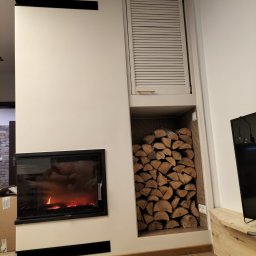 Suchodolski - Nowoczesny kominek z czarną obudową i paleniskiem, obok nisza na drewno opalanego drewnem, zamknięta szafka z żaluzjami, drewniana podłoga i fragment telewizora.