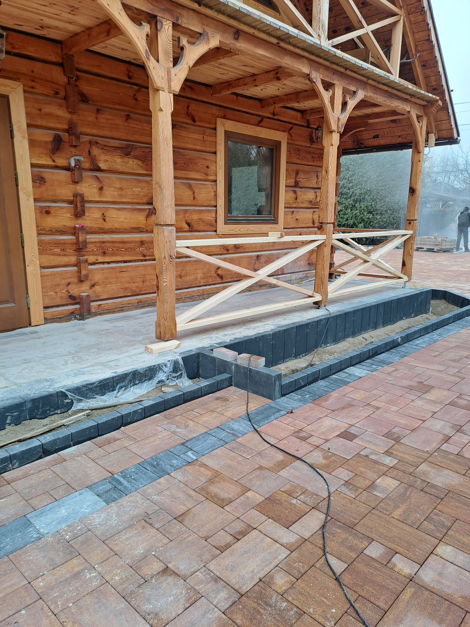Drewniany dom z bali w trakcie budowy tarasu z kostki brukowej. Widoczne elementy konstrukcyjne domu i częściowo ułożona kostka w odcieniach brązu i szarości.