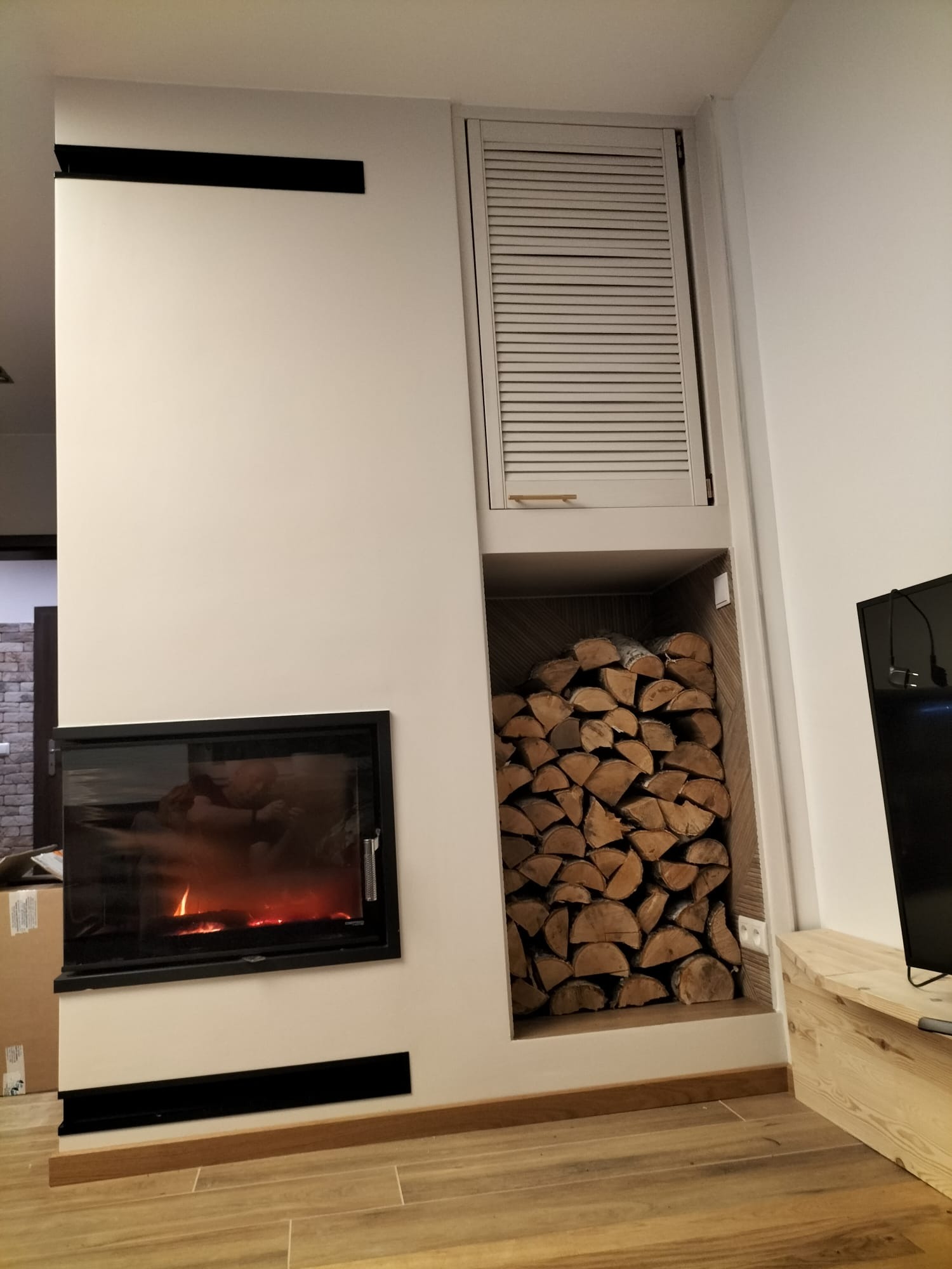 Nowoczesny kominek z czarną obudową i paleniskiem, obok nisza na drewno opalanego drewnem, zamknięta szafka z żaluzjami, drewniana podłoga i fragment telewizora.