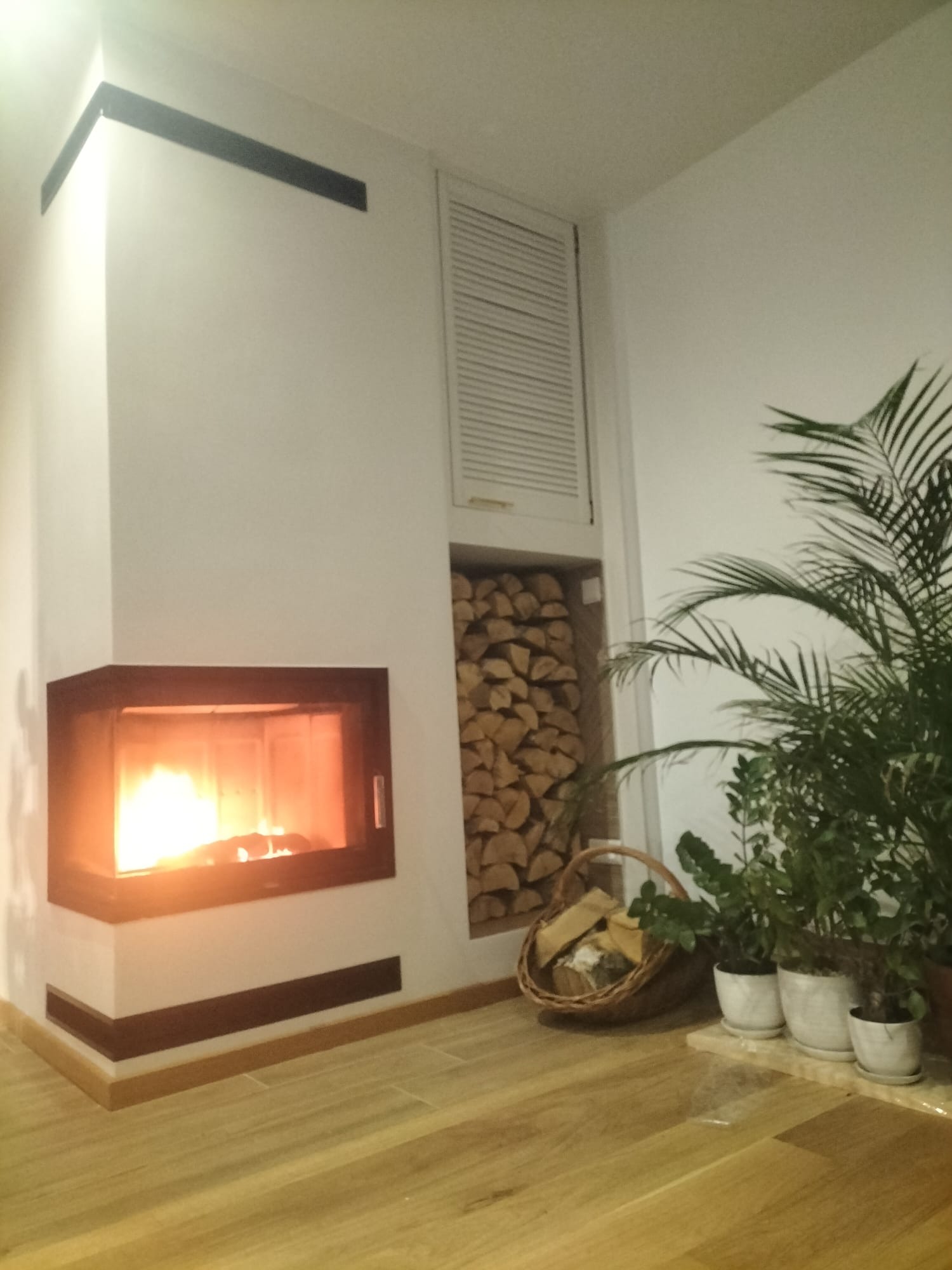 Nowoczesny kominek narożny z paleniskiem, obok drewno ułożone w niszy ściennej, rośliny doniczkowe i drewniana podłoga. Minimalistyczny design wnętrza.
