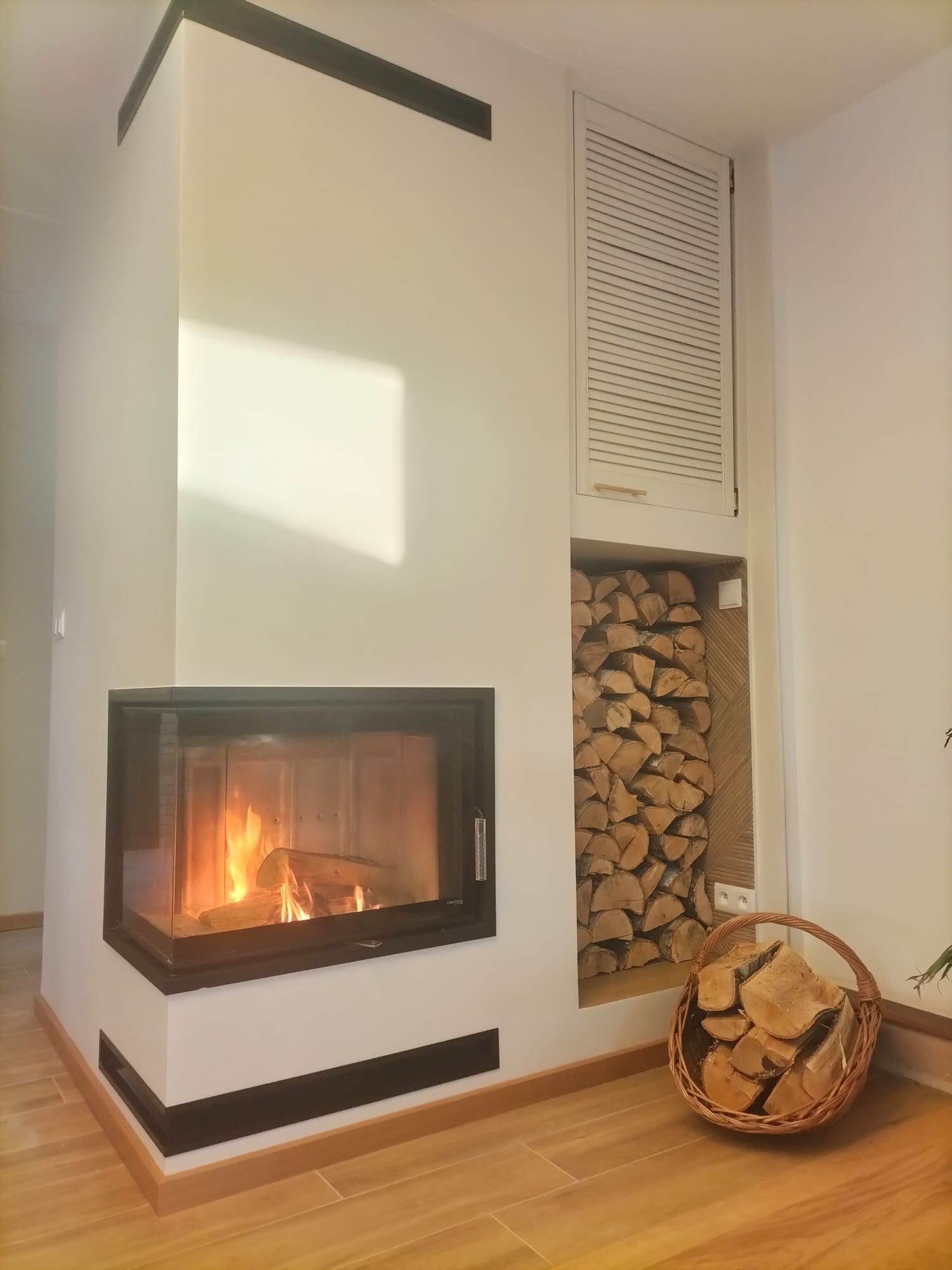 Nowoczesny kominek z paleniskiem i zapasem drewna w ścianie, kosz z drewnem obok. Jasne wnętrze z drewnianą podłogą. Minimalistyczny design.