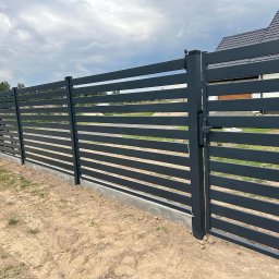 Spawik Bauservice Daniel Zatwarnicki - Nowoczesne, poziome ogrodzenie i furtka z metalowych profili pomalowanych na grafitowo, widoczne na tle domu i pochmurnego nieba.