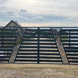 Spawik Bauservice Daniel Zatwarnicki - Nowoczesna, pozioma brama wjazdowa w kolorze grafitowym z geometrycznym wzorem trójkątów, widok z zewnątrz posesji, dom w tle.