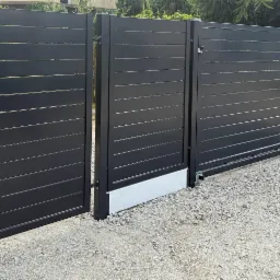 Nowoczesna, przesuwna brama wjazdowa w kolorze antracytowym z poziomymi panelami. Częściowo otwarta furtka z zamkiem. Szara nawierzchnia z kamyków, tło z zielenią.