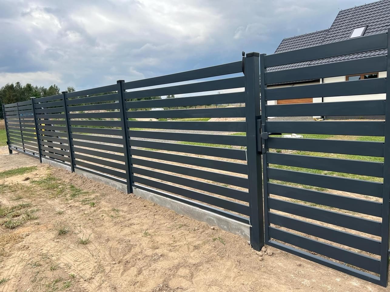 Nowoczesne, poziome ogrodzenie i furtka z metalowych profili pomalowanych na grafitowo, widoczne na tle domu i pochmurnego nieba.