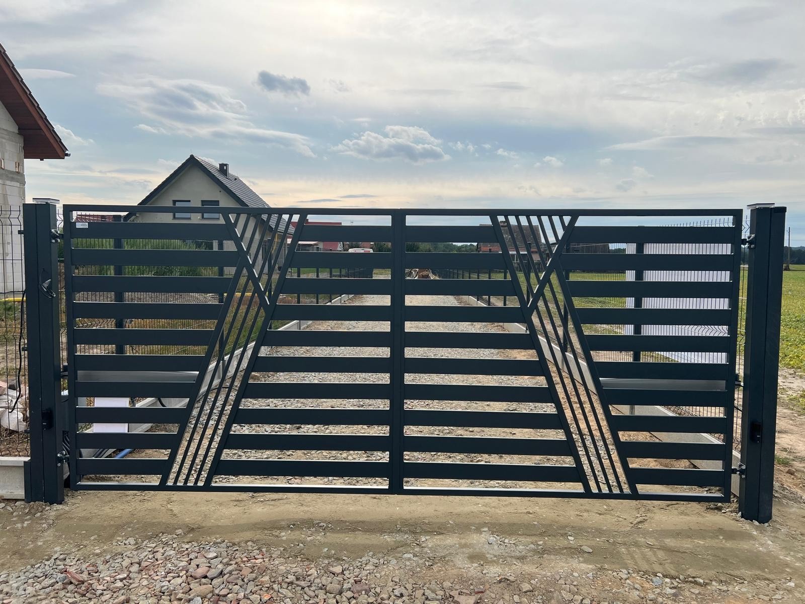 Nowoczesna, pozioma brama wjazdowa w kolorze grafitowym z geometrycznym wzorem trójkątów, widok z zewnątrz posesji, dom w tle.