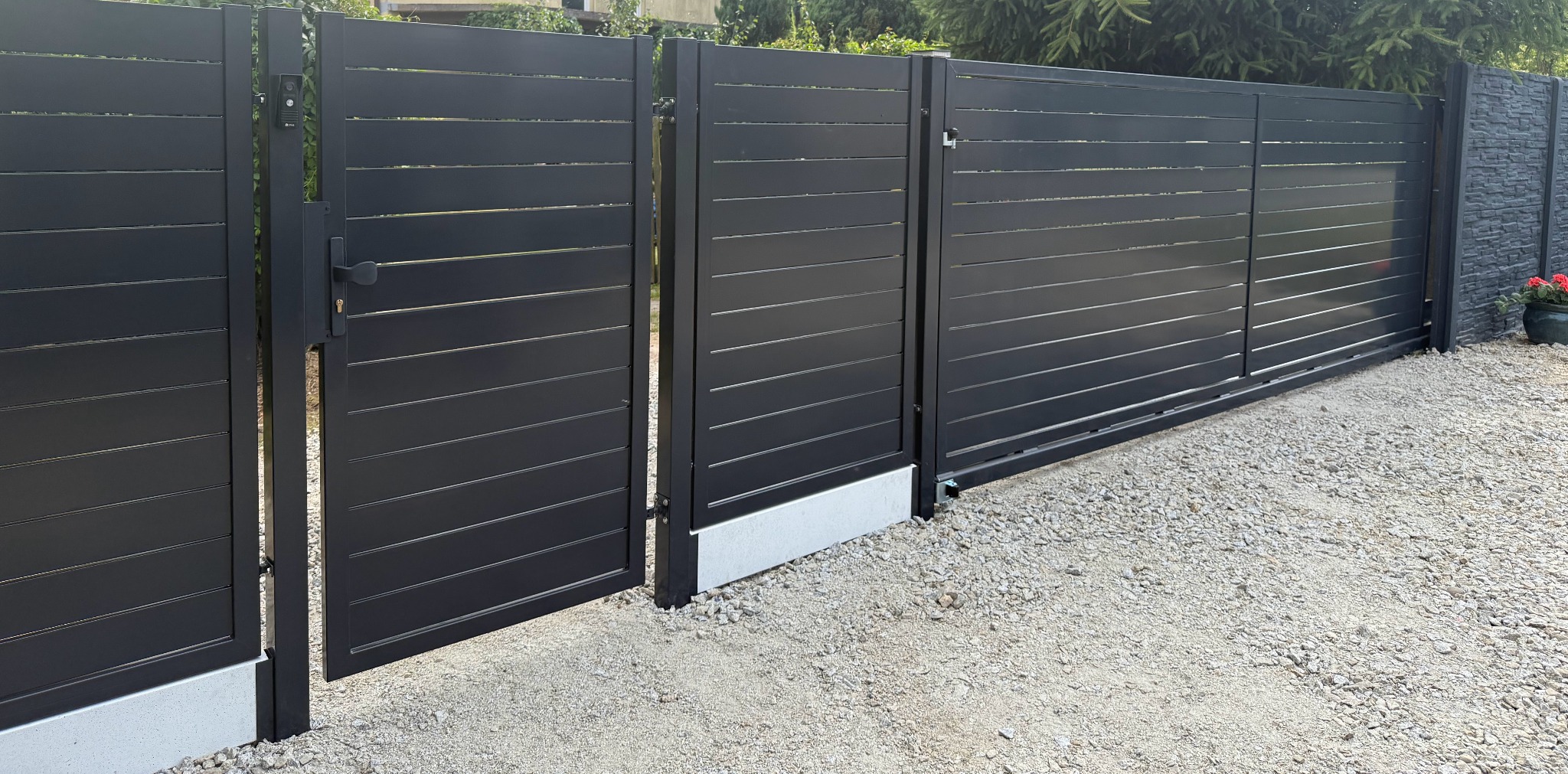 Nowoczesna, przesuwna brama wjazdowa w kolorze antracytowym z poziomymi panelami. Częściowo otwarta furtka z zamkiem. Szara nawierzchnia z kamyków, tło z zielenią.
