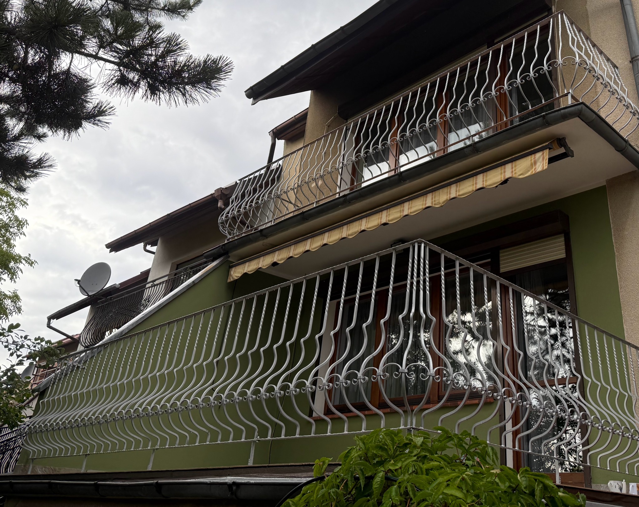 Dwukondygnacyjny dom z balkonami zabezpieczonymi kutymi, ozdobnymi balustradami. Widok z dołu, fragment drzewa w lewym górnym rogu kadru.