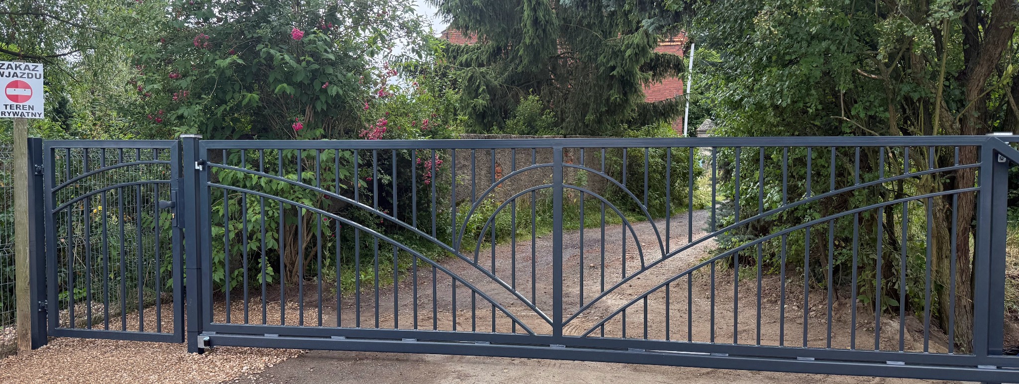 Metalowa brama wjazdowa przesuwna w kolorze grafitowym, z ażurowym wzorem, otoczona zielenią i znakiem zakazu wjazdu. Widok z zewnątrz posesji.