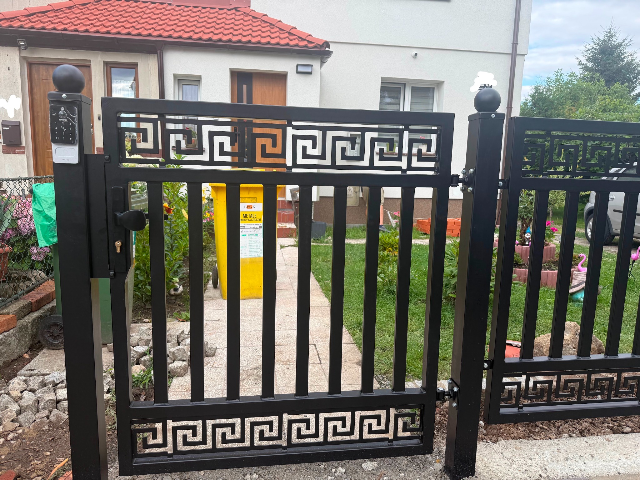 Czarna, metalowa furtka z dekoracyjnym wzorem greckim, z domofonem na słupku, prowadząca do domu z czerwonym dachem. Widok z bliska, z żółtym pojemnikiem na śmieci w tle.