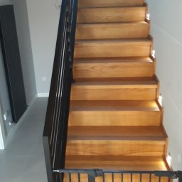 PPUH JAR DREW - Drewniane schody z czarną balustradą i oświetleniem LED w stopniach, zabezpieczone bramką dla dzieci. Widok z dołu, nowoczesny design.