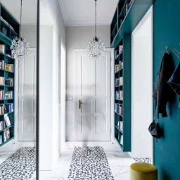 PPUH JAR DREW - Korytarz z zabudową biblioteczną w kolorze morskim, lustrem i wzorzystą posadzką. Kryształowy żyrandol i dwa pufy dopełniają aranżację.