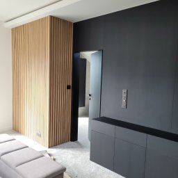 PPUH JAR DREW - Nowoczesne wnętrze z drewnianą ścianką działową z lameli, czarną zabudową meblową i szarą sofą. Minimalistyczny design z dbałością o detale i oświetlenie.