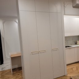 PPUH JAR DREW - Biała szafa wnękowa z dekoracyjnymi frezami i złotymi uchwytami, wkomponowana w aranżację mieszkania w Buku. Widoczna zabudowa kuchenna w tle.