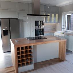 PPUH JAR DREW - Jasnoszara kuchnia na wymiar z drewnianym blatem wyspy, płyta indukcyjna na wyspie oraz regał na wino, widoczna zabudowa piekarnika i lodówka side-by-side.