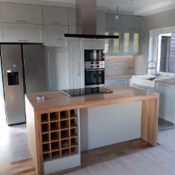 Jasnoszara kuchnia na wymiar z drewnianym blatem wyspy, płyta indukcyjna na wyspie oraz regał na wino, widoczna zabudowa piekarnika i lodówka side-by-side.