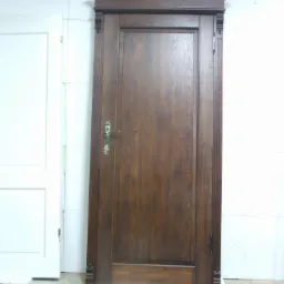 Ciemnobrązowe drzwi wewnętrzne z ozdobnymi listwami i złotą klamką, stojące obok białych drzwi na tle białej ściany w trakcie montażu.