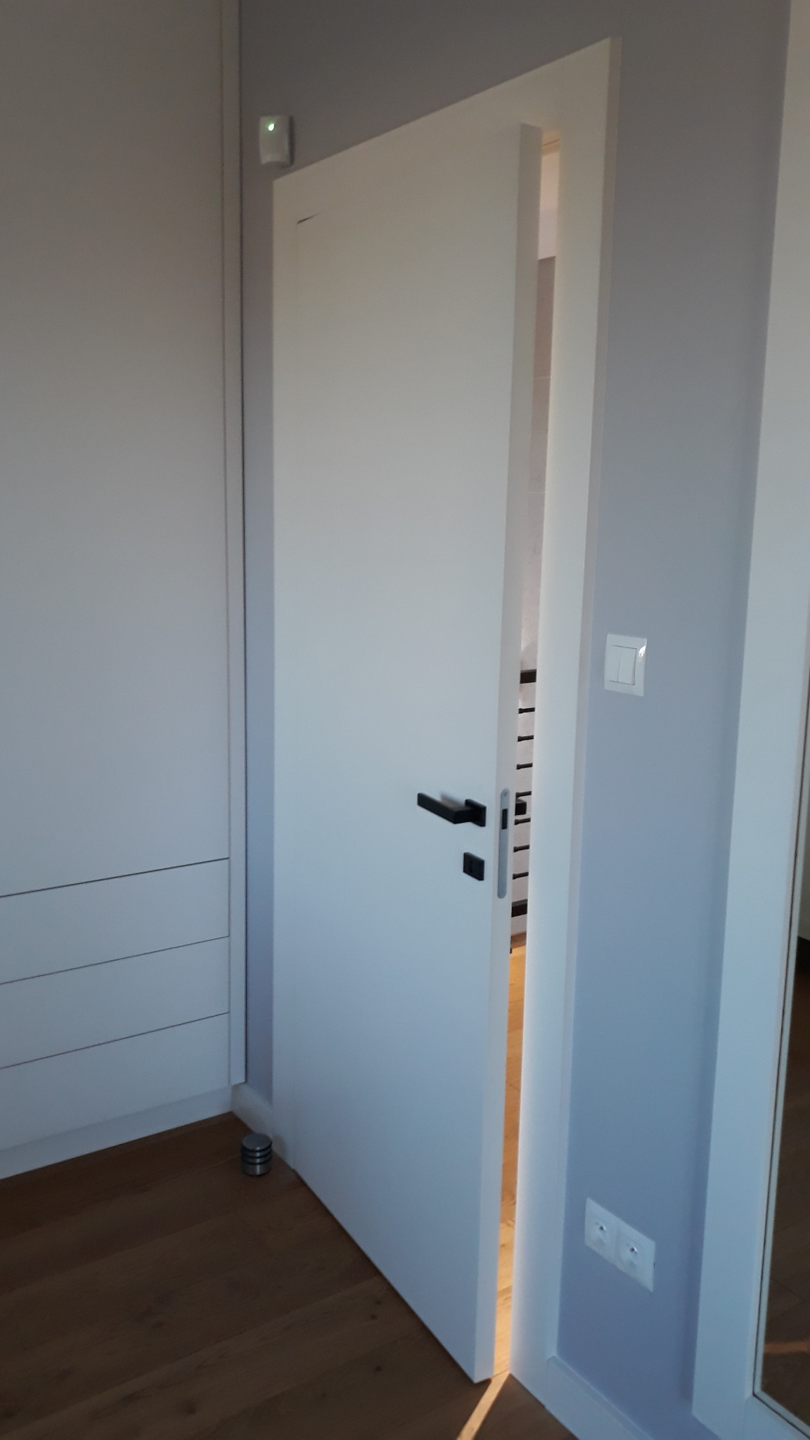 Białe, minimalistyczne skrzydło uchylone, czarny, prosty uchwyt, widok na schody w głębi. Nowoczesne wykończenie, drewniana podłoga, szara ściana z włącznikiem.